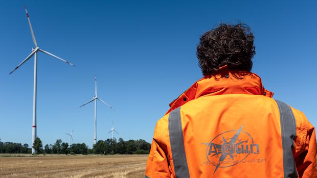 Een inspecteur van A-Quin, gekleed in een oranje jas, kijkt naar meerdere windmolens in een open veld onder een heldere blauwe lucht.