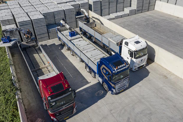 Luchtfoto van drie vrachtwagens die betonnen platen laden op een groot opslagterrein, omgeven door gestapelde betonnen platen